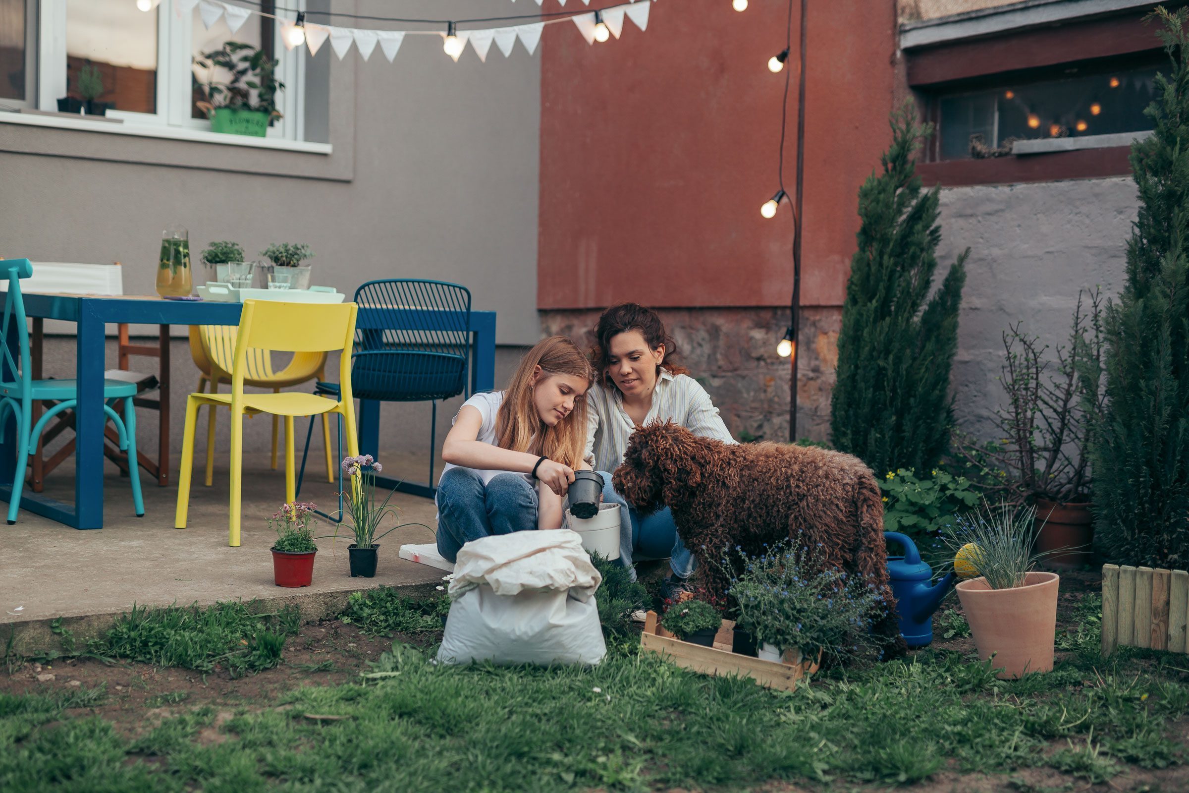 Zwei Frauen sitzen vor ihrem Eigenheim auf dem Gras und pflanzen um, während ein Hund zuschaut.
