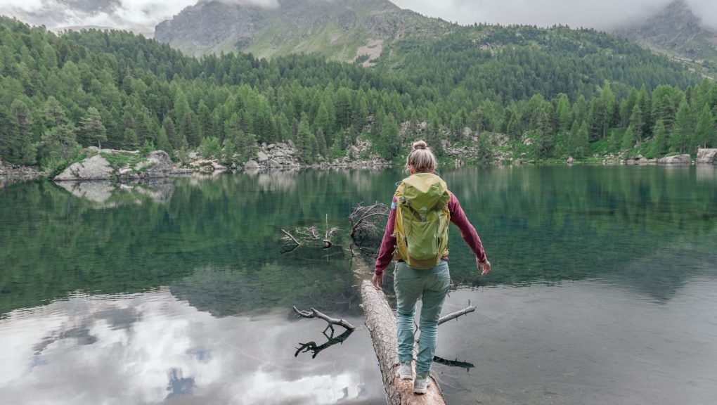 Eine Frau mit Rucksack überquert einen See auf einem Baumstamm.