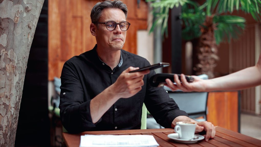 Ein Mann sitzt an einem Tisch mit einer Tasse Kaffee und bezahlt mit Mobilepayment.